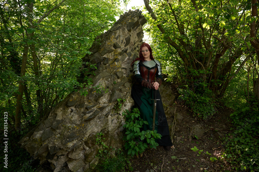 Fototapeta premium Mittelalterliche Frau mit Rüstung und roten Haaren als Kriegerin mit Kriegsbemalung mit Schwert im Sommer vor einer alten Burgruine, mit Korsett aus Leder und Umhang