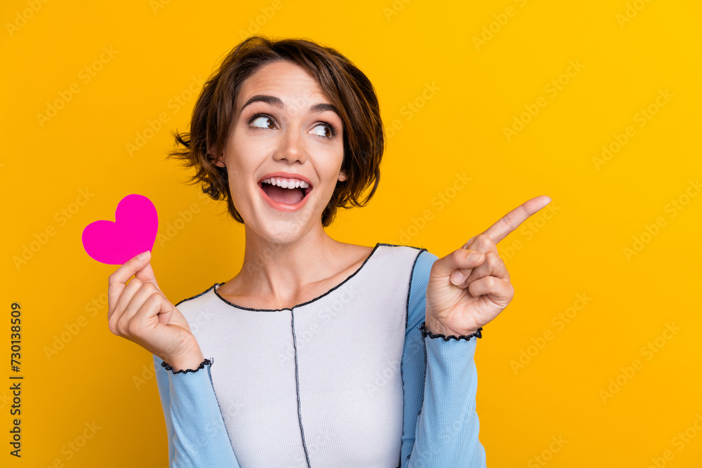 Photo of adorable excited woman wear top holding pink heart looking pointing empty space isolated yellow color background Photo of adorable excited woman wear top holding pink heart looking pointing empty space isolated yellow color background