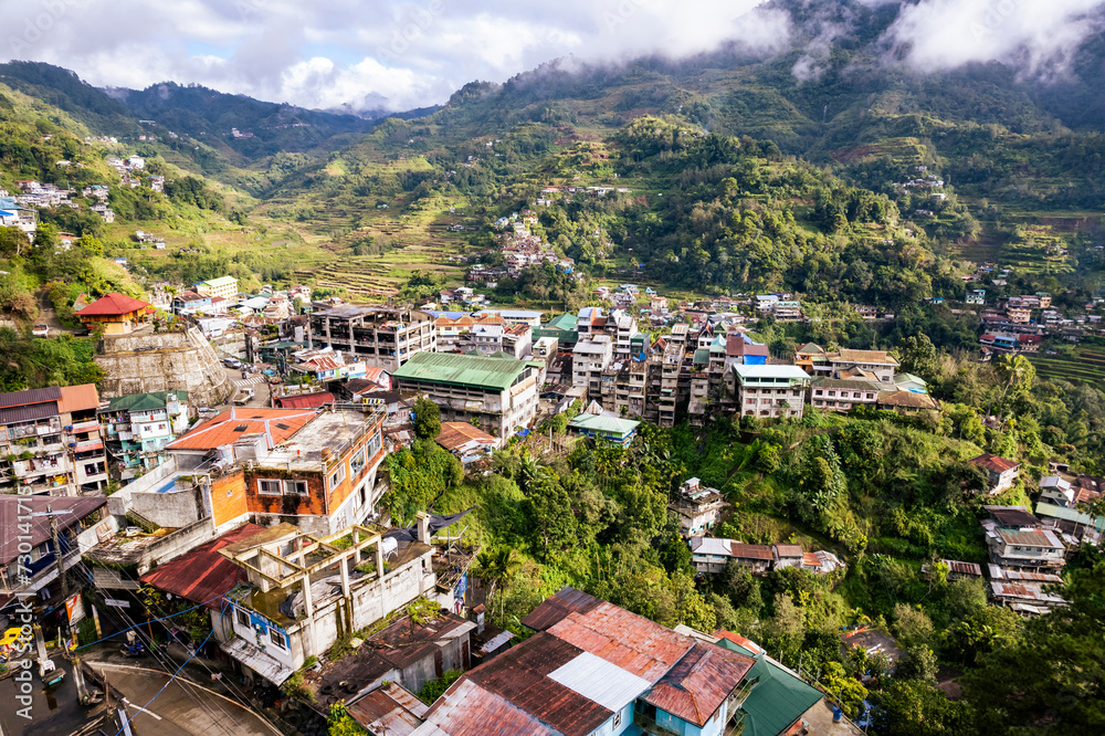 Aerial view of a mountainous town with lush greenery and typical hillside houses, enveloped in morning mist. At the town of Banaue, in the province of Ifugao, Philippines.