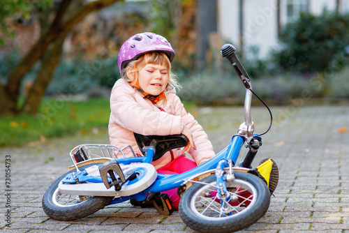Fototapeta Naklejka Na Ścianę i Meble -  Cute little girl sitting on the ground after falling off her bike. Upset crying preschool child with safe helmet getting hurt while riding a bicycle. Active family leisure with kids.