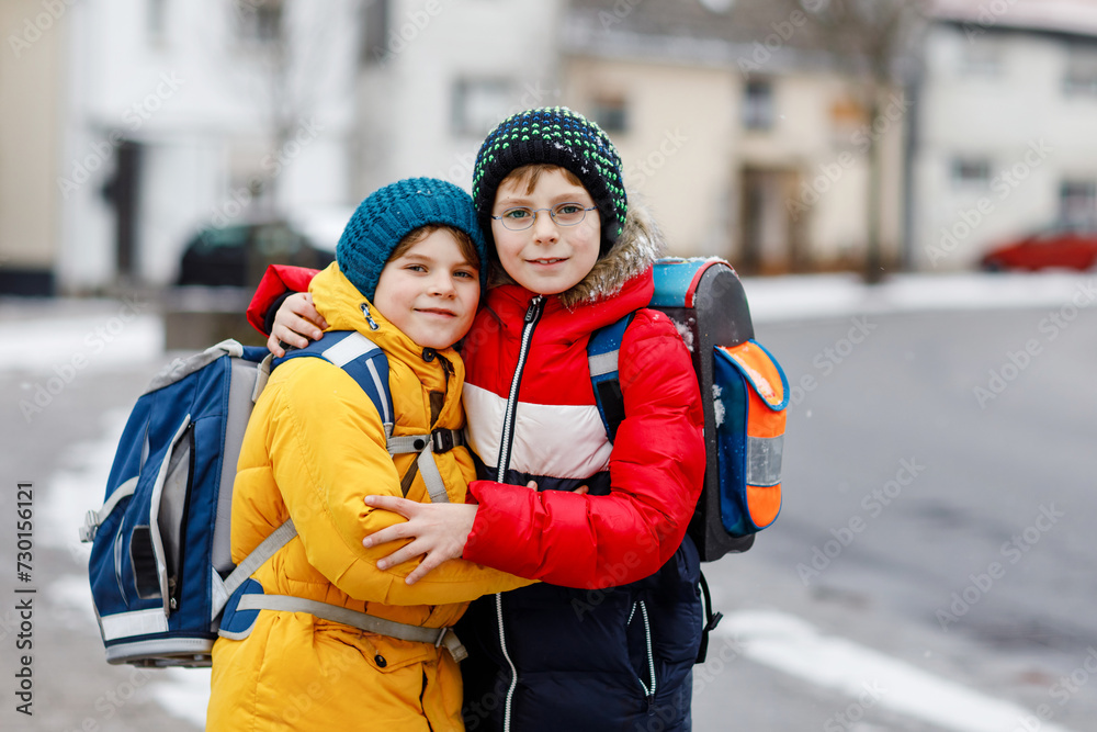 Two little kids boys of elementary class walking to school during snowfall. Happy children having fun and playing with first snow. Siblings and best friends with backpack in colorful winter clothes. Two little kids boys of elementary class walking to school during snowfall. Happy children having fun and playing with first snow. Siblings and best friends with backpack in colorful winter clothes.