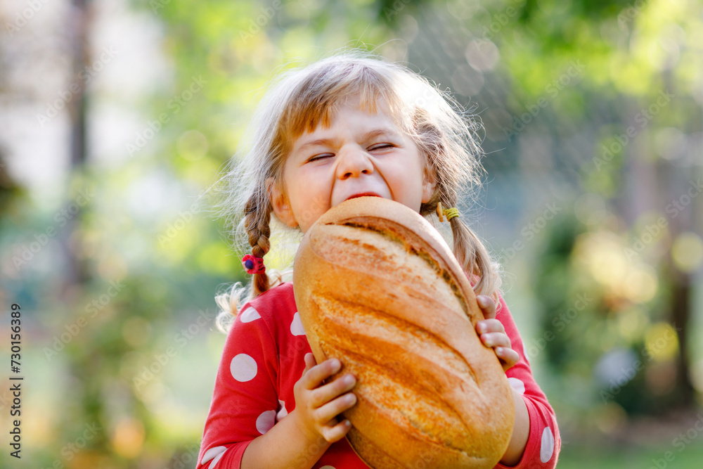 Little toddler girl holding big loaf of bread. Funny happy child biting and eating healthy bread, outdoors. Hungry kid.