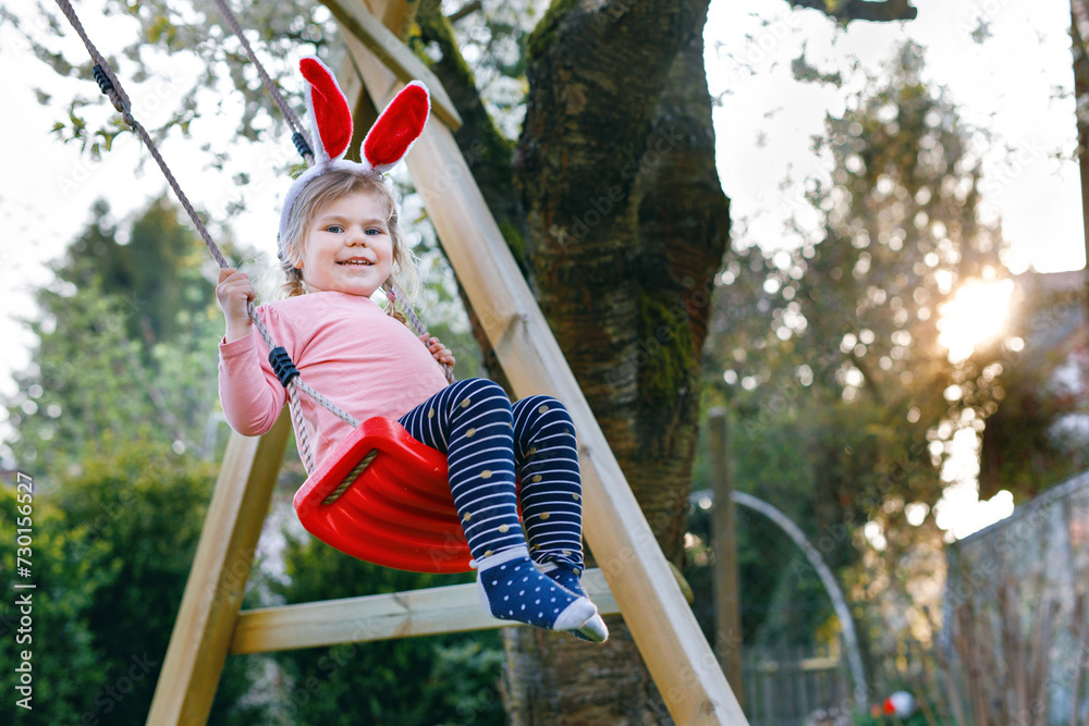 Happy beautiful little toddler girl with red Easter bunny ears having fun on swing in domestic garden. Cute healthy child swinging under blooming trees on sunny spring day. Baby laughing and crying