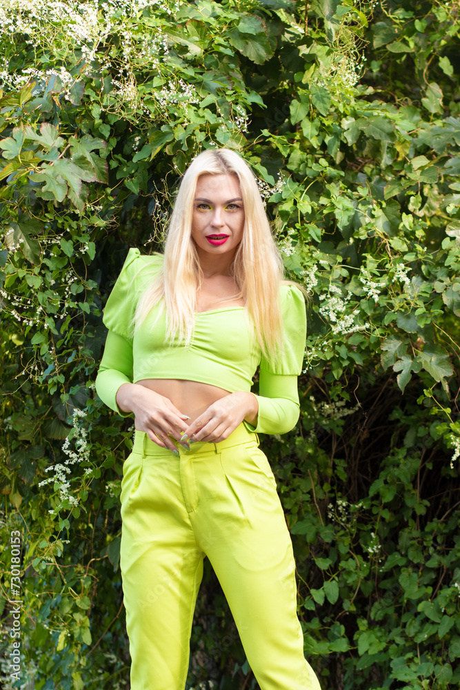 Portrait of a beautiful young blonde with long hair in a yellow-green pantsuit against a background of green bushes with flowers Portrait of a beautiful young blonde with long hair in a yellow-green pantsuit against a background of green bushes with flowers