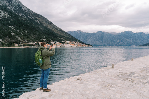 Man with a backpack stands on a pier by the sea and photographs the mountains. Back view