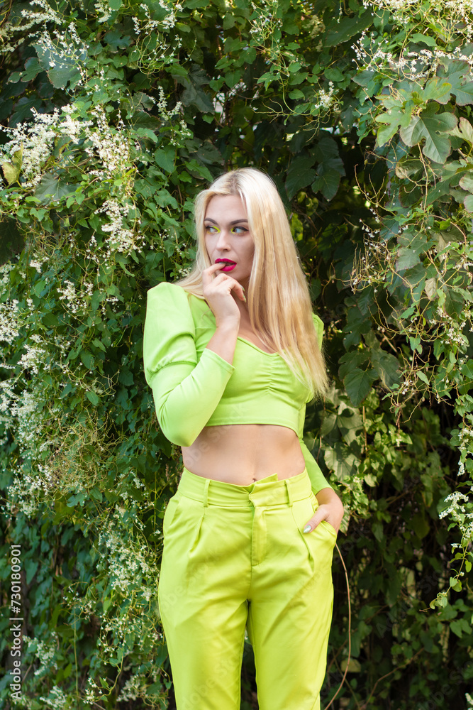Portrait of a beautiful young blonde with long hair in a yellow-green pantsuit against a background of green bushes with flowers Portrait of a beautiful young blonde with long hair in a yellow-green pantsuit against a background of green bushes with flowers