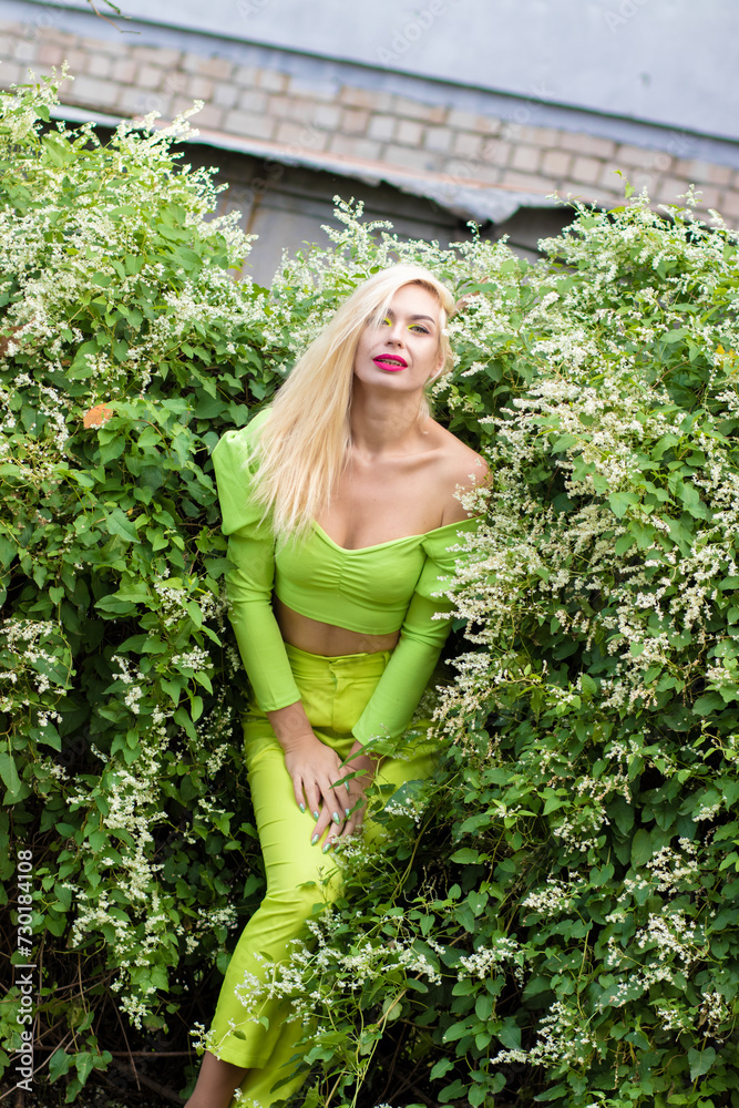 Full-length portrait of a beautiful young blonde with long hair in a yellow-green pantsuit and bright high-heeled pumps against a background of green bushes with flowers Full-length portrait of a beautiful young blonde with long hair in a yellow-green pantsuit and bright high-heeled pumps against a background of green bushes with flowers