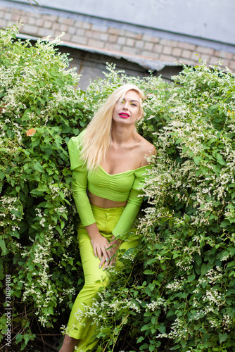 Full-length portrait of a beautiful young blonde with long hair in a yellow-green pantsuit and bright high-heeled pumps against a background of green bushes with flowers