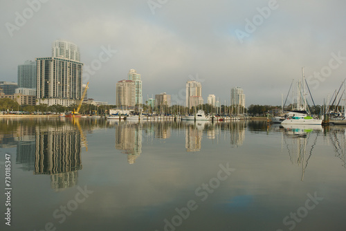 Wide shot Looking north from Demens Landing Park over smooth water with reflections towards city scape of St. Petersburg, Florida .  Sailbots in marina with Cloudy blue and white sky with light fog.  © Del Harper