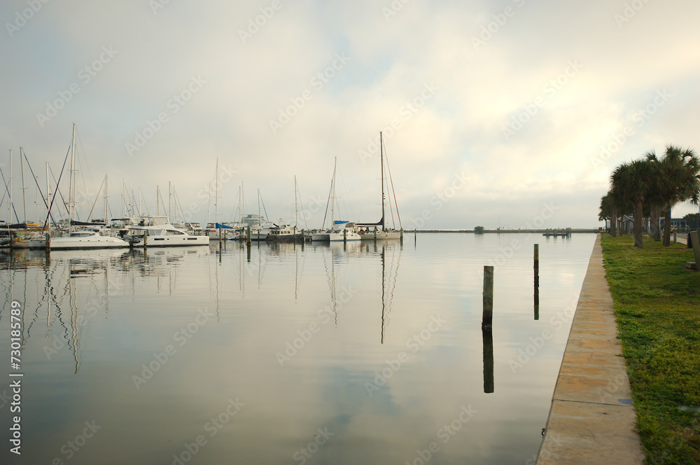 Obraz premium View east leading lines seawall at South Yacht Basin in St. Petersburg, FL. Green Palm Trees on right and boat marina on the left. Airport in the background early AM cloudy after fog lifts.