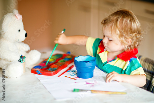 Cute adorable baby girl learning painting with water colors. Little toddler child drawing at home  using colorful brushes. Healthy happy daughter experimenting with colors  water at home or nursery.