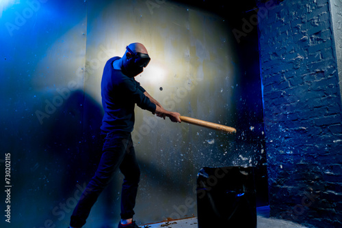 A man in a protective mask smashes a glass bottle with a bat in a crash room for stress relief