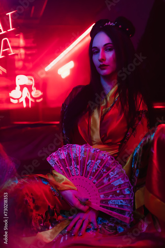 Close-up of a beautiful girl in a kimono sits on a red sofa with a fan in her hand. The image of a geisha, Chinese national costume. Party concept, night club