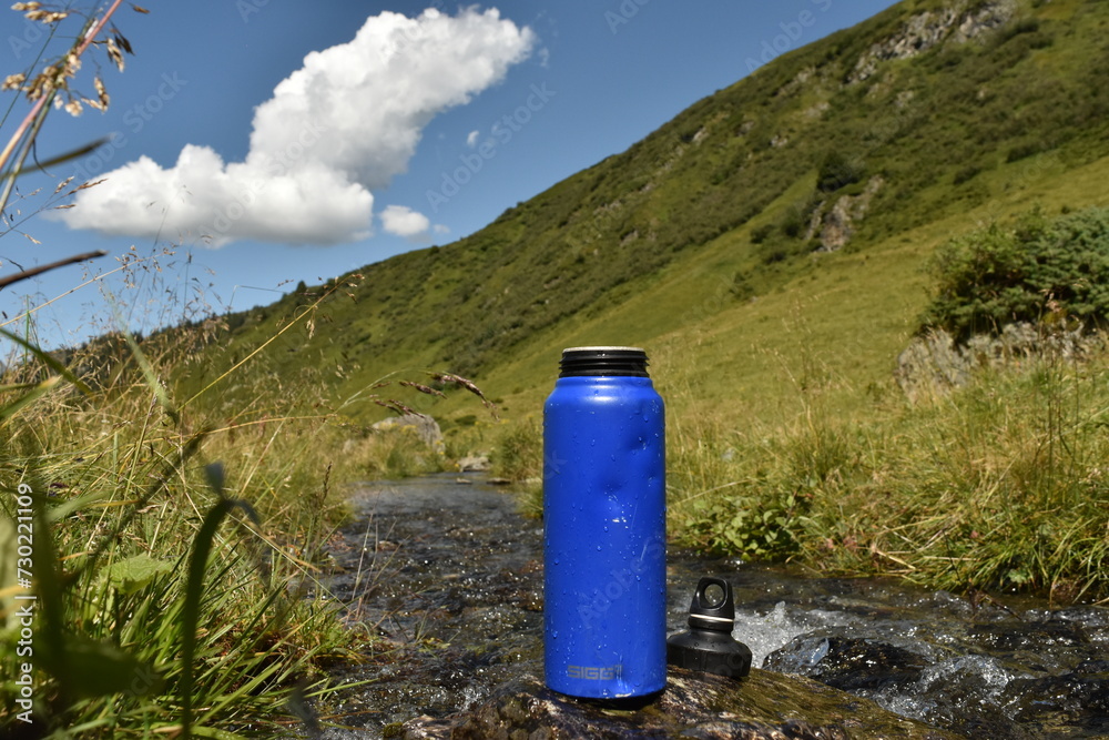 Obraz premium Blue Reusable Water Bottle by Mountain Stream in Sunny Hillside Environment
