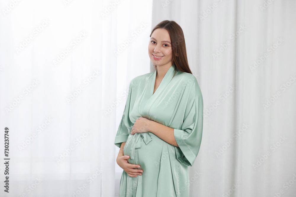 Beautiful pregnant woman in green dressing gown near window indoors Beautiful pregnant woman in green dressing gown near window indoors