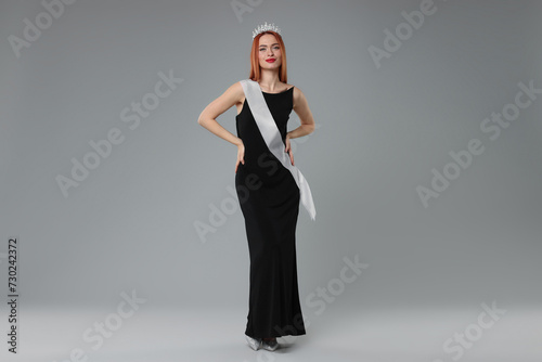 Beautiful young woman with tiara and ribbon in dress on light grey background. Beauty contest