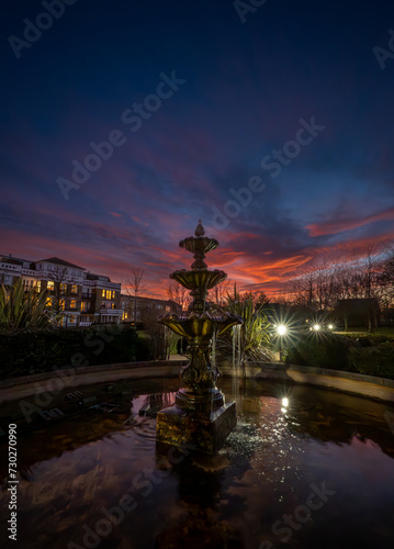 Fountain and pond in a park at sunset with a pink sky. Night view in portrait orientation with buildings and trees behind.