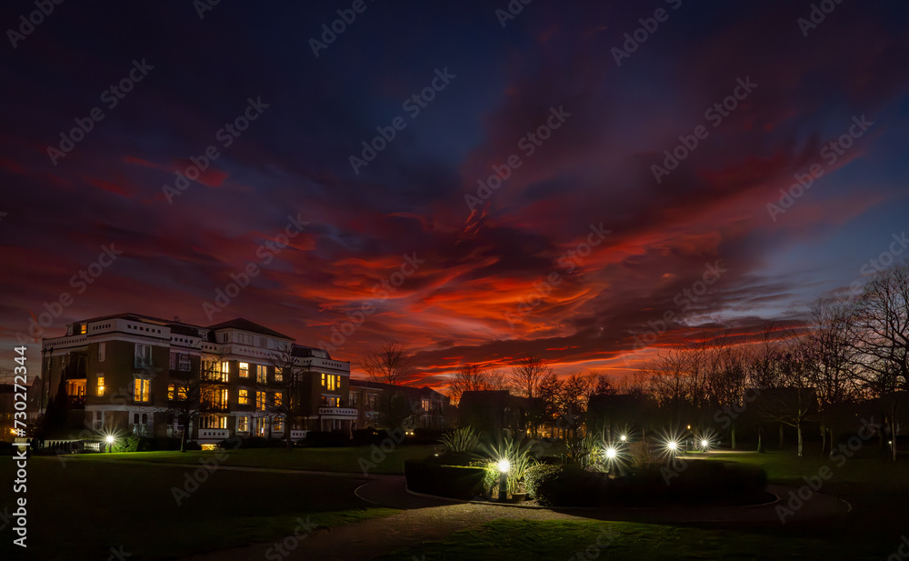 Park with lights at sunset with a fiery red sky. Night view in landscape orientation with buildings and trees behind.