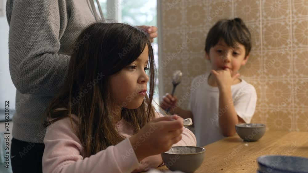 Pensive little girl eating yogurt from bowl in kitchen with mother and little brother in the background. Domestic authentic lifestyle scene of family eating breakfast early in the morning Pensive little girl eating yogurt from bowl in kitchen with mother and little brother in the background. Domestic authentic lifestyle scene of family eating breakfast early in the morning
