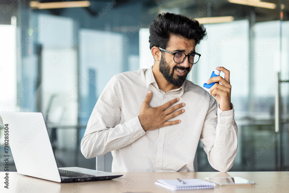 Overworked indian businessman feeling unwell at office desk with laptop and inhaler Overworked indian businessman feeling unwell at office desk with laptop and inhaler