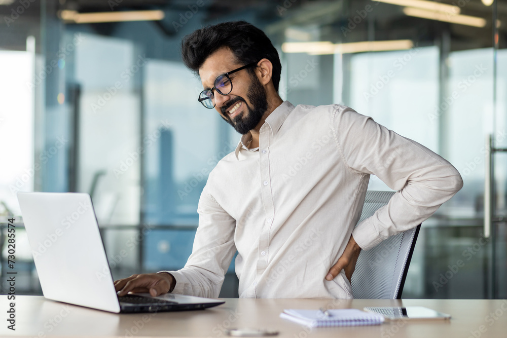 Exhausted indian businessman with back pain working late on laptop at office desk Exhausted indian businessman with back pain working late on laptop at office desk
