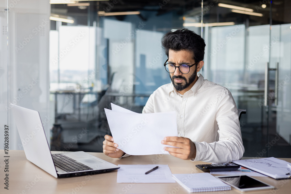 Focused indian businessman analyzing documents at office desk with laptop and smartphone Focused indian businessman analyzing documents at office desk with laptop and smartphone