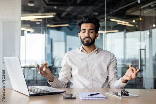 Indian businessman meditating at office desk with laptop for mindfulness