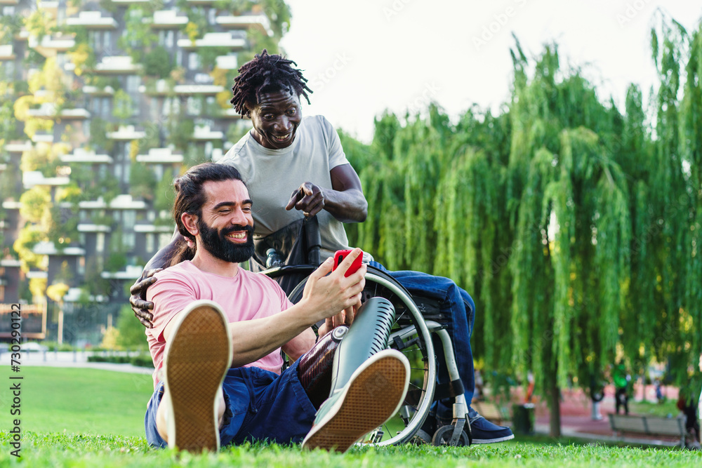 Obraz premium Joyful friends, one with a prosthetic leg and the other in a wheelchair, sharing a laugh in a sunny park setting.