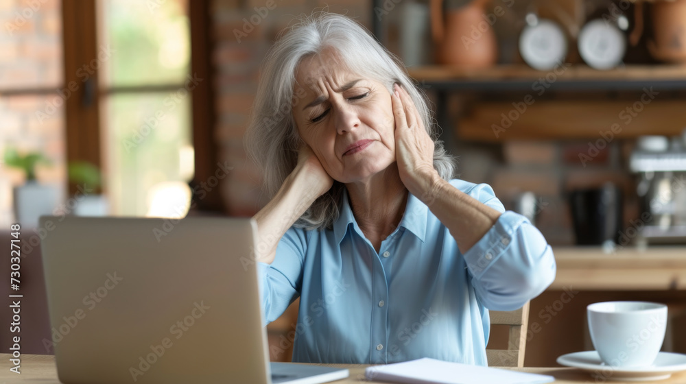 An elderly woman is sitting at a table with a laptop, holding her head in pain or stress. An elderly woman is sitting at a table with a laptop, holding her head in pain or stress.
