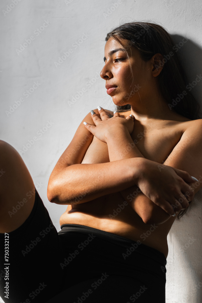 Fototapeta premium brunette latina woman covering her breasts with her hands folded while leaning against the wall with a sad look looking down.