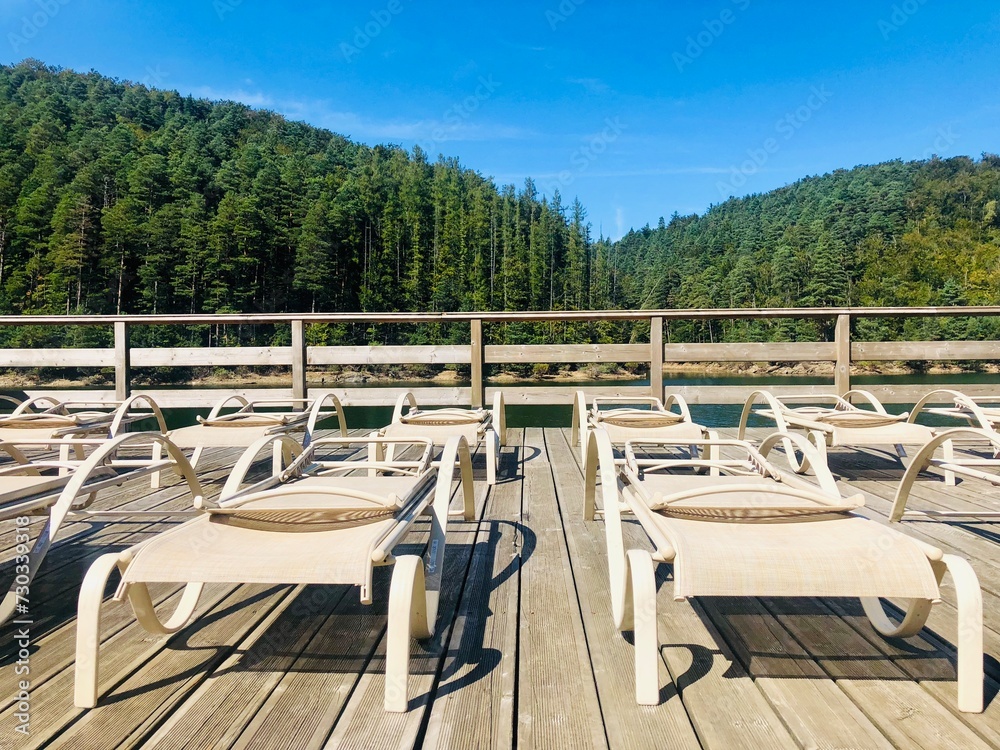 Fototapeta premium Empty sunbeds on a wooden pontoon near a lake surrounded by green forest