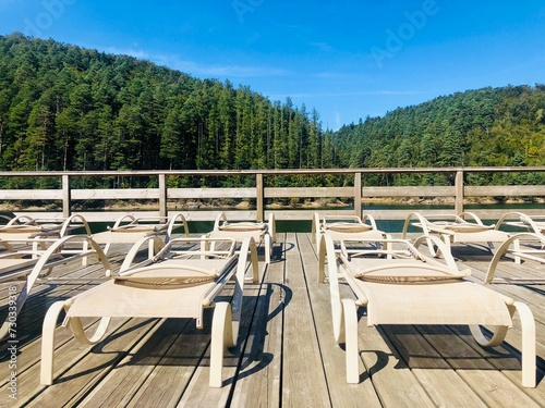 Empty sunbeds on a wooden pontoon near a lake surrounded by green forest