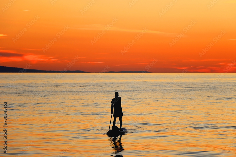 Unrecognizable person is stand up paddleboarding on the beach at sunset. Beautiful landscape in Brela, Croatia.