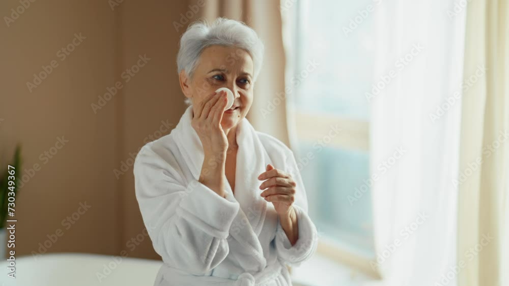 Elderly woman touching face, look at mirror. Healthy smiling old woman in bathrobe with gray hair taking care of her face at home. Getting old age change, face treatment beauty self-care concept. Elderly woman touching face, look at mirror. Healthy smiling old woman in bathrobe with gray hair taking care of her face at home. Getting old age change, face treatment beauty self-care concept.