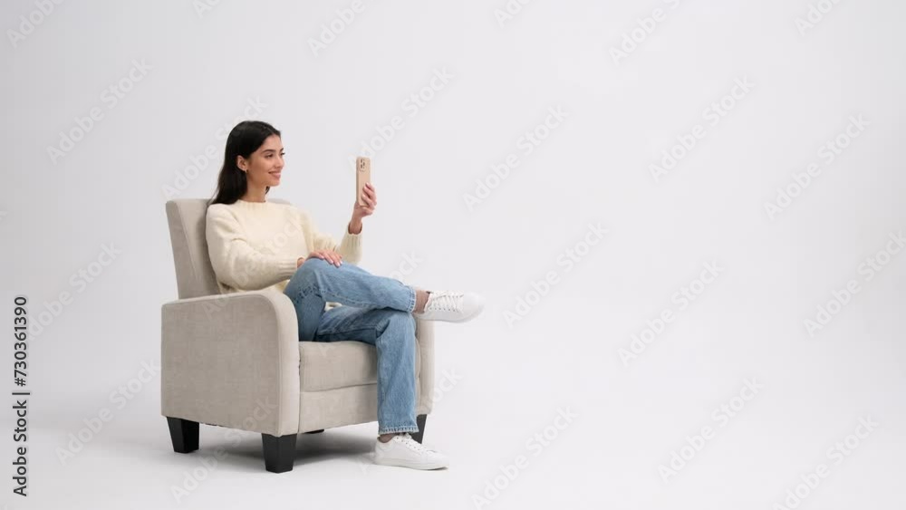Active Caucasian woman talking on a video call and waving hand in greeting using the smartphone in a soft comfortable armchair on a white background. Video conference, video call with friends concept. Active Caucasian woman talking on a video call and waving hand in greeting using the smartphone in a soft comfortable armchair on a white background. Video conference, video call with friends concept.