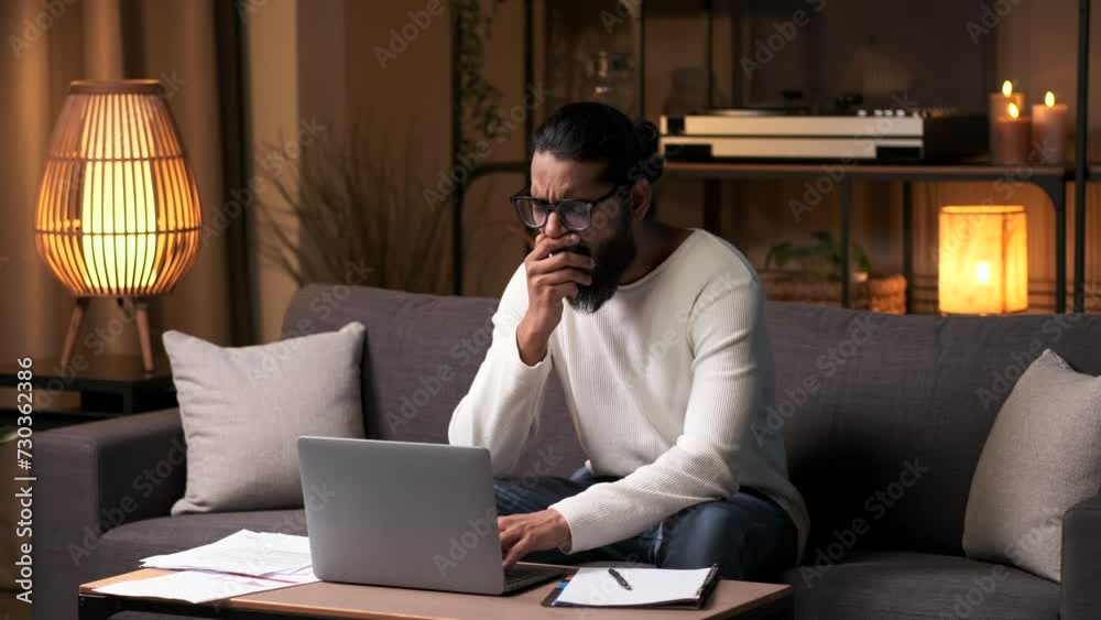 Tired and sleepy Indian male businessman works on a laptop at home in the evening and yawns, feeling tired from late work, overtime, deadlines, stress. Tired and sleepy Indian male businessman works on a laptop at home in the evening and yawns, feeling tired from late work, overtime, deadlines, stress.