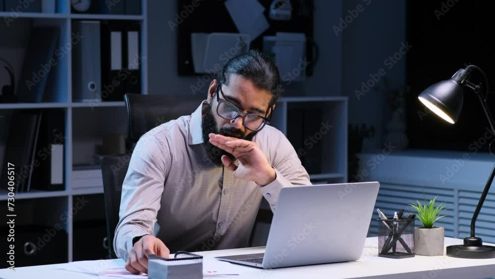 Overworked Indian businessman working late with documents and yawning in the evening office. High workload, stress and burnout, working before deadline.