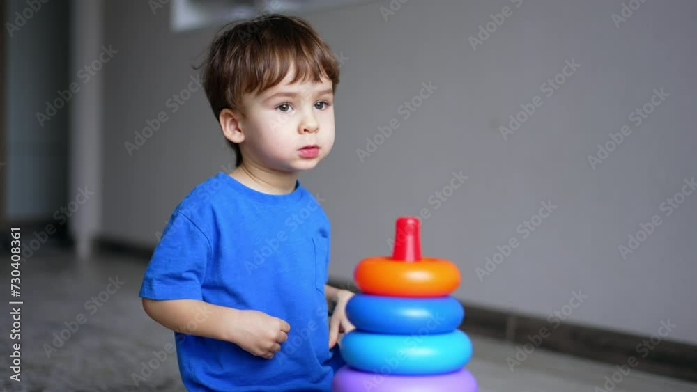 Caucasian toddler in blue t-shirt plays with toy pyramid. Kid looks ahead and points at something with his finger. Caucasian toddler in blue t-shirt plays with toy pyramid. Kid looks ahead and points at something with his finger.