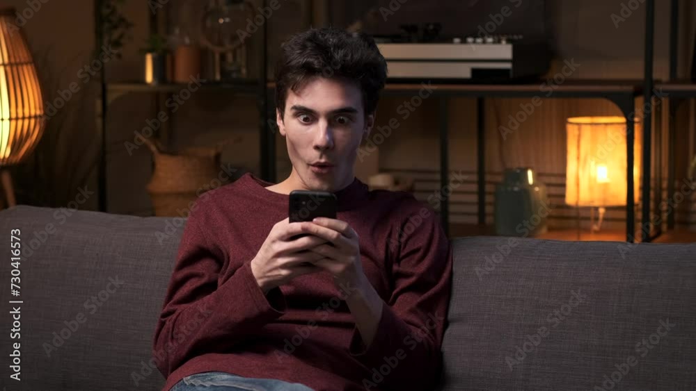 Young Caucasian man is surprised when texting on mobile phone and exclaiming wow while relaxing on the sofa in the living room in the evening. Online communication, internet entertainment. Young Caucasian man is surprised when texting on mobile phone and exclaiming wow while relaxing on the sofa in the living room in the evening. Online communication, internet entertainment.