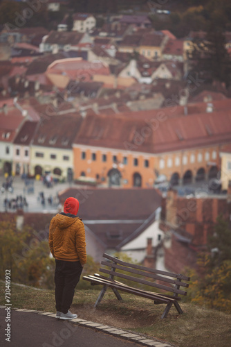 View from back. Young man enjoying the view of the city on an autumn day. A traveler man with red hood enjoys the view to the cityscape of Brasov, Romania. Amazing scenic view of the old town.