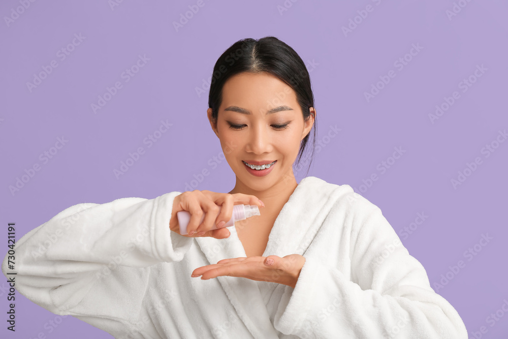 Young Asian woman in bathrobe applying serum on lilac background Young Asian woman in bathrobe applying serum on lilac background
