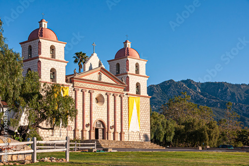 Santa Barbara, CA, USA - April 20, 2009: Old Mission church front facade in sun  under blue sky. Green trees and mountains in back. The Santa Barbara Mission was established on the Feast of Saint Barb