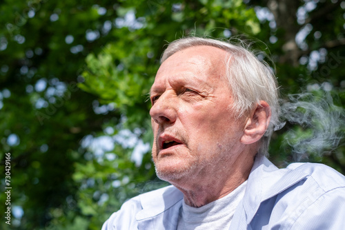 Old man smoke of smoking in the air in a sunlit park, candid shot suitable for discussions about lifestyle and aging, unhealthy habits and addictions, insurance ad
