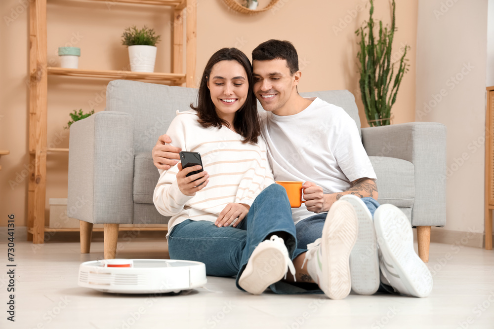Young couple using mobile phone for remote control of modern robot vacuum cleaner in living room Young couple using mobile phone for remote control of modern robot vacuum cleaner in living room