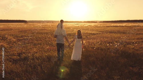Father with lovely daughter perched on shoulders strolls through field with alongside wife. Father walks hand in hand with wife. Father with little girl on shoulders and wife enjoy walk in meadow photo