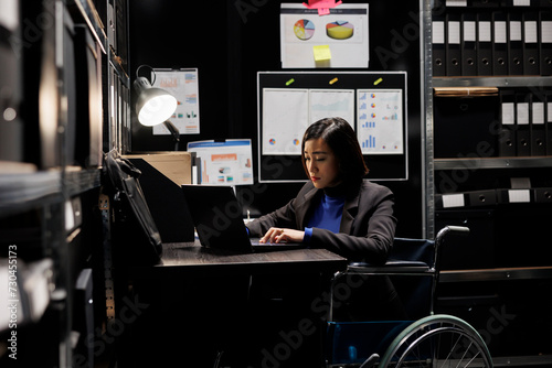 Professional businesswoman accountancy employee in wheelchair writing budget plan and accountancy strategy on laptop. Asian woman in repository filled with document folders and flowcharts