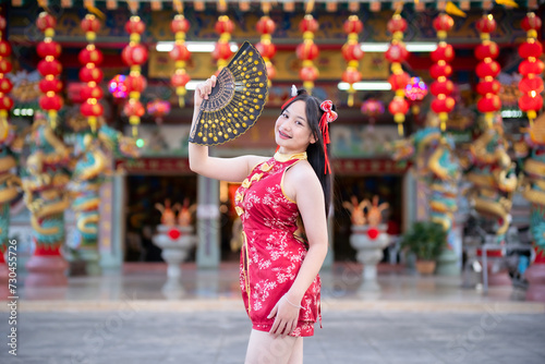 Portrait beautiful cute smiles Asian young woman wearing red traditional Chinese cheongsam decoration and holding a Chinese Fanning for Chinese New Year Festival at Chinese shrine in Thailand