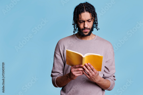 Concentrated arab man reading paperback book with yellow cover. Focused young person with thoughtful expression studying literature, holding softcover textbook on blue background photo