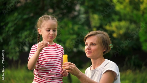 Happy mother with joyful daughter blows bubbles in park among trees on vacation. Preschooler girl waits till mother blows bubbles in meadow at weekend. Mother and child spend time blowing bubbles