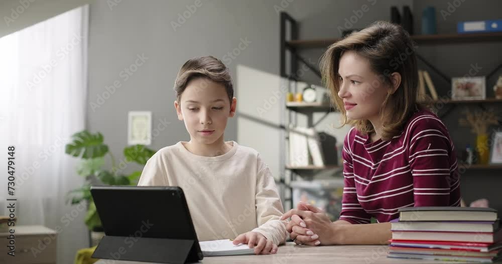 Mother helping son doing homework at home desk, studying online with digital tablet. Private tutor teaches preteen boy individually Mother helping son doing homework at home desk, studying online with digital tablet. Private tutor teaches preteen boy individually